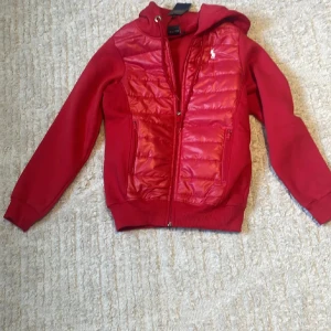 Röd pufferjacka från Ralph Lauren - Riktigt snygg röd pufferjacka från Ralph Lauren med huva och dragkedja. Jackan har quiltade paneler framtill i glansigt material och klassisk logga broderad på bröstet. Ej äkta då jag köpte den i spanien för ca 6 månader sedan för 1500kr💕