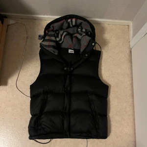 Burberry väst/jacka  - Säljer en svart dunväst från Burberry med avtagbara ärmar och huva. Västen har klassiskt rutigt foder i grått, rött och vitt samt dragkedja och snörning. Perfekt för lager-på-lager och riktigt snygg streetstil.
