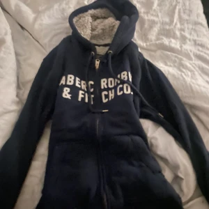 Mörkblå teddyjacka Abercrombie & Fitch - Mysig mörkblå teddyjacka från Abercrombie & Fitch med vit text på bröstet. Jackan har huva, dragkedja och fluffigt teddyfoder i hela insidan. Perfekt för kalla dagar när du vill vara både varm och snygg. Klassisk college-vibe och skön passform. Pris kan diskuteras!