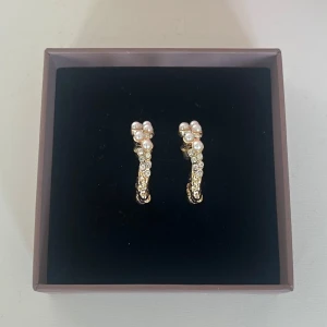 Guldiga örhängen med pärlor och stenar - Snygga guldiga örhängen i halvbåge med vita pärlor och glittrande stenar. De har en lyxig känsla och passar perfekt om du vill sticka ut lite extra. Örhängena är lätta att bära och ger en elegant touch till din stil.