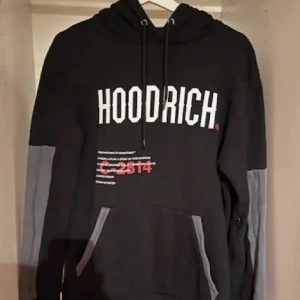 Svart och grå HOODRICH hoodie - Svart hoodie från HOODRICH med stora vita bokstäver på bröstet och tryck i rött och vitt. Grå detaljer på ärmar och ficka, samt snörning i huvan. Perfekt för en streetwear-look och riktigt skön passform. Byxor finns