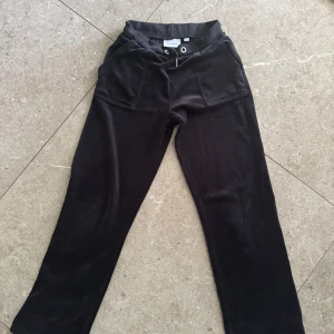 Svarta mjukisbyxor Juicy Couture - Svarta mjukisbyxor från Juicy Couture med dragsko i midjan, stora fickor fram och bak med snygga metallknappar och broderad JC-logga. Byxorna är i mjuk velour och har en relaxed passform som är både chill och trendig.