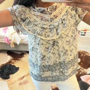 Blommig volangblus med spetsdetaljer - Supersöt blus i vitt med blått blommönster och spetsdetaljer. Volang vid axlar och bröst, knappar framtill och korta ärmar. Tunn och luftig, perfekt för vår och sommar. 💕💕💖💗💞😻😽😸