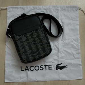 Svart axelväska Lacoste - Snygg svart axelväska från Lacoste med geometriskt vitt mönster och diskret Lacoste-logga framtill. Väskan har justerbar axelrem och är tillverkad i tåligt syntetmaterial. Perfekt storlek för mobil, plånbok och andra essentials.