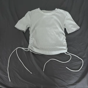 Ljusgrå ribbad topp från Zara - Supersnygg ljusgrå ribbad topp från Zara med korta ärmar och cool snörning i sidorna som gör att du kan justera passformen. Mjuk och stretchig i materialet, perfekt till jeans eller kjol. Enkel men trendig design med rund hals.