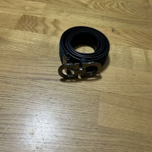 Svart skärp med metallspänne Ferragamo - Snyggt svart bälte från Ferragamo med ett ikoniskt metallspänne i silverfärg. Bältet är smalt och stilrent, tillverkat i skinn med en diskret textur. Perfekt accessoar för att lyfta din outfit med en lyxig touch.