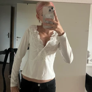 Vit topp med spetsdetaljer - Supersöt vit topp med v-ringning och breda spetsdetaljer längs urringningen. Långärmad och lätt croppad passform som ger en feminin vibe. Perfekt att styla med jeans eller kjol för en trendig look. Jättebra skick och i storlek 158/164 men passar xs!! 