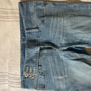 Ljusblå bootcut jeans med slitningar - Säljer ett par ljusblå jeans med bootcut-modell och slitna detaljer framtill samt fransade benslut. Byxorna har låg midja, klassiska fem fickor och stängs med tre knappar.