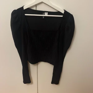 Svart långärmad topp från H&M Divided - Säljer en svart långärmad topp från H&M Divided i storlek S. Toppen har en croppad passform, fyrkantig urringning och puffiga axlar. Materialet är plisserat och ger en cool struktur. Perfekt att styla med höga jeans eller kjol för en trendig look.