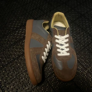 Gats  - Snygga sneakers i en mix av brunt mocka och blått skinn, med vita snören och gummisula. Klassisk retrostil med rund tå och låg profil. Perfekta för dig som gillar stilrena och tidlösa sneakers med en twist. Helt nya andvända 1 gång. Storlek 43 men passar bättre på folk som har storlek 42. Obs. Det är inte maison margellas 