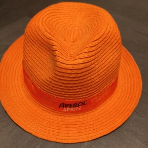 Orange Aperol Spritz hatt - Snygg orange hatt och solglasögon från Aperol Spritz. Hatten har klassisk form och dekorativt band runt kronan där loggan är tryckt. Hatten är tillverkad i ett flätat syntetmaterial och har en bred brätte som ger cool sommarvibe.