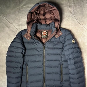 Colmar dunjacka - Säljer en mörkblå dunjacka från Colmar med brunt foder och avtagbar huva. quiltad design och Colmar-logga på ärmen. Perfekt för kyliga dagar och riktigt snygg passform. Storlek 50/M