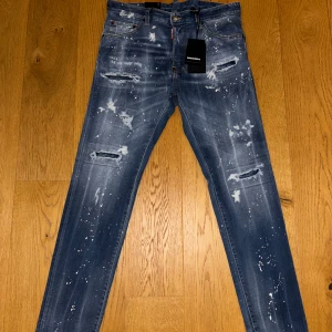 Helt nya! Dsquared2 blå jeans med slitningar - Snygga blå jeans från Dsquared2 med coola slitningar och vit färgstänk över hela byxan. Modellen har klassisk femficksdesign, smal passform och detaljerade rivna partier framtill. Perfekt för dig som gillar streetstyle och vill sticka ut.