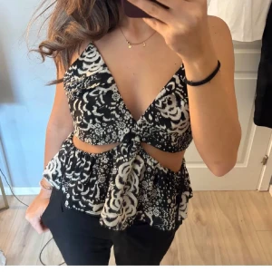 Svart och vit omlottopp från Zara - Säljer en snygg omlottopp från Zara i svart och vitt med blommigt mönster. Toppen har djup v-ringning, volang nedtill och cut-out detaljer vid midjan. Axelbandslös framtill med smala axelband bak. Perfekt för dig som gillar trendiga och unika plagg!