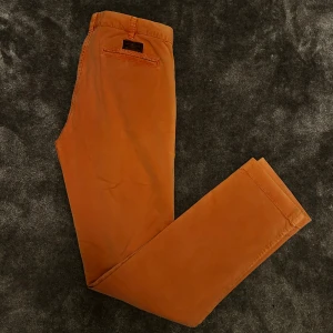 Orange chinos från Morris, 32/33 - Snygga orange chinos från Morris i storlek 32/33. Byxorna är tillverkade i mjuk bomull med lite stretch för extra komfort. Klassisk rak passform med fickor bak och diskret logga ovanför ena bakfickan. Perfekta för en stilren och avslappnad look.
