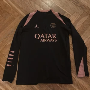 PSG x Jordan långärmad träningströja - Säljer en svart långärmad Paris Saint-Germain träningströja från Jordan med rosa detaljer och Qatar Airways-tryck. Tröjan har halv dragkedja, Dri-Fit-material och rosa text på ärmen. Perfekt för fotbollsträning eller streetwear.