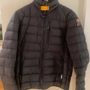 Svart pufferjacka från Parajumpers - Svart pufferjacka från Parajumpers med klassisk gul detalj vid kragen och patch på ärmen. Jackan har quiltad design, tre dragkedjefickor och hög krage. Perfekt för kalla dagar och har en clean, sportig look som funkar till streetwear.