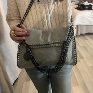 Grå axelväska med kedjedetaljer. Stella McCartney liknande  - Snygg grå Stella McCartney liknande väska i mjuk mocka med mörka silverfärgade kedjor längs kanterna och som axelrem. Väskan dekorativa sömmar runtom. 💞