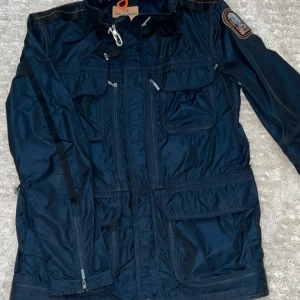 Mörkblå vindjacka Parajumpers M - Snygg mörkblå vindjacka från Parajumpers med flera fickor, dragkedjor och cool patch på ärmen. Jackan har meshfoder för ventilation och orange detaljer på insidan. Perfekt för dig som gillar funktion och stil i ett.