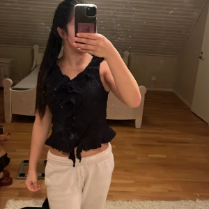 Svart volangblus från Gina Tricot - Supersnygg svart blus från Gina Tricot med volangdetaljer framtill och lätt skrynklig struktur. Ärmlös modell med v-ringning och knyt längst ner. Perfekt för dig som gillar trendiga och feminina toppar. på näst skita bilden så är de två jätte små håll som int syns ett dugg