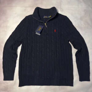 Marinblå stickad tröja Polo Ralph Lauren - Kabelstickad marinblå tröja från Polo Ralph Lauren med halv dragkedja och hög krage. Tröjan har lång ärm, ribbade muddar och en röd broderad logga på bröstet. Perfekt för kyliga dagar och snygg till jeans.