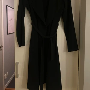 Svart trenchcoat med bälte - Säljer en svart trenchcoat med lång passform och bälte i midjan. Jackan har långa ärmar och breda slag framtill, vilket ger en clean och stilren look. Perfekt för dig som gillar minimalistisk stil och vill ha en jacka som funkar till många outfits.