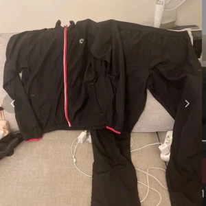 Svart vindjacka från Clique Sports L - Svart vindjacka från Clique Sports i storlek L. Jackan har en röd och vit dragkedja framtill, röda detaljer vid ärmslut och krage samt två sidofickor med dragkedja. Materialet är lätt och syntetiskt, perfekt för sportiga dagar. Snygg och enkel design med hög krage.