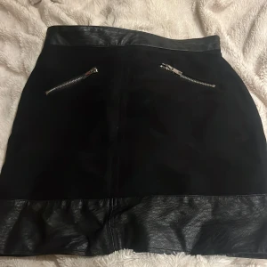 Svart kortkjol med skinn och dragkedjor - Snygg svart kortkjol med detaljer i skinn upptill och nedtill. Kjolen har två silvriga dragkedjor framtill som ger en cool vibe. Perfekt för dig som gillar en edgy stil och vill sticka ut lite extra.