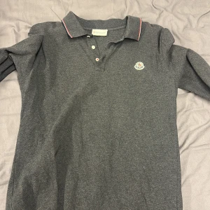 Mörkgrå Moncler pikétröja långärmad - Snygg mörkgrå långärmad pikétröja från Moncler med klassisk krage och knappar framtill. Tröjan har Moncler-logga på bröstet och diskreta röda och vita detaljer på krage och ärmslut. Perfekt för dig som gillar stilrena och exklusiva plagg.