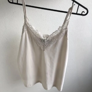 Beige/vit linnetopp med spetsdetaljer - Supersöt Vit linne med tunna axelband som går att justera och v-ringning dekorerad med spets. Köpt här på Plick men tjejen skrev den va vit och på hennes bild såg den vit ut men den va mer Beige än vit i verkligheten