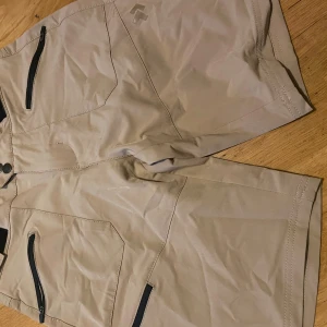 Beiga shorts från Black Diamond - Säljer ett par beiga shorts från Black Diamond i storlek small. De har flera praktiska fickor med dragkedja framtill och ficklock bak. Materialet är lätt och syntetiskt, perfekt för aktiva dagar. Justerbar midja med kardborre för bästa passform.