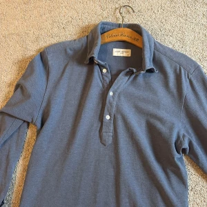 Blå långärmad pike från John Henric - Snygg blå långärmad pike från John Henric med klassisk krage och tre knappar framtill. Skjortan är långärmad och tillverkad i mjuk bomull, perfekt för en avslappnad men stilren look.