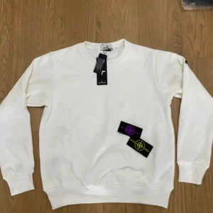 Vit sweatshirt från Stone Island - Vit sweatshirt från Stone Island med klassisk rund hals och ribbade muddar. Medföljer två utbytbara Stone Island badges i lila och gul. Tröjan har en clean look och är tillverkad i mjuk bomull, perfekt för dig som gillar streetwear och vill ha något stilrent.