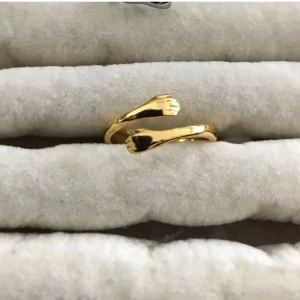 Guldfärgad ring med handdetaljer - Unik guldfärgad ring med två små händer som möts framtill. Ringen har en öppen design och glänsande finish som ger en cool och personlig touch. Perfekt för dig som gillar annorlunda smycken med roliga detaljer.