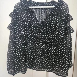 Svart prickig blus med volanger - Säljer en svart blus från Mango med vita prickar och volangdetaljer längs axlar och bröst. Blusen har v-ringad hals och långa ärmar i ett lätt, transparent material som ger en luftig känsla. Perfekt för dig som gillar trendiga mönster och detaljer.