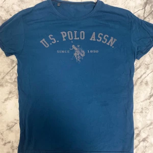 Blå t-shirt från U.S. Polo Assn. - Snygg blå t-shirt från U.S. Polo Assn. med tryck på bröstet och klassisk rund hals. T-shirten har korta ärmar och är gjord i mjuk bomull, perfekt för en chill och sportig look. Trycket visar polospelare och texten 'Since 1890'.