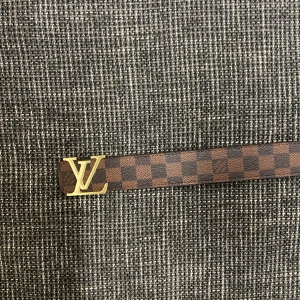 Louis Vuitton rutigt brunt bälte - Säljer ett Louis Vuitton bälte i klassiskt brunt och mörkbrunt rutigt mönster med ikoniskt LV-spänne i guld. Bältet är gjort i skinn och har en exklusiv känsla, perfekt för att lyfta vilken outfit som helst.