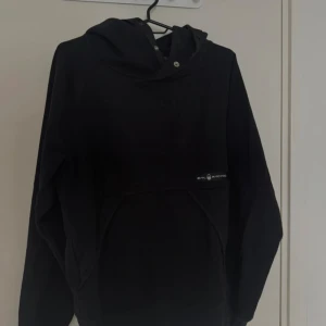 Svart hoodie från Sail Racing - Svart hoodie från Sail Racing med huva och snygga tryckknappar vid halsen. Tröjan har en klassisk känguruficka framtill och diskret logga på bröstet. Perfekt för dig som gillar stilren och sportig look.