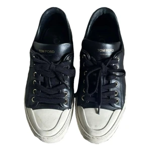 Svarta sneakers från Tom Ford - Snygga svarta sneakers från Tom Ford med vit platt sula och guldfärgade detaljer. Skorna är i skinn och har snörning framtill samt rund tå. Logotypen 'TF' i guld syns på hälen och 'Tom Ford' på plösen. Perfekt för dig som gillar stilrena och lyxiga sneakers.