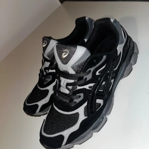 Svarta och grå Asics Gel sneakers - Säljer ett par Asics Gel sneakers i svart och grått med coola detaljer och mesh-paneler för extra ventilation. Skorna har snörning och en chunky platt sula som ger skön komfort. Perfekt för dig som gillar sportig stil och vill ha något som sticker ut.