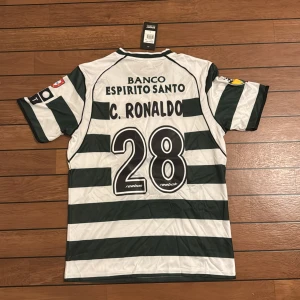 Sporting CP C. Ronaldo fotbollströja L - Säljer en officiell Sporting CP fotbollströja från Reebok i storlek L, med C. Ronaldo och nummer 28 på ryggen. Tröjan har breda gröna och vita ränder, klubbmärke på bröstet och sponsorer på ärmarna. Tillverkad i lätt polyester, perfekt för match eller samling.