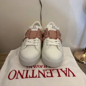 Valentino Rockstud sneakers vit/rosa - Snygga Valentino Rockstud sneakers i vitt skinn med ljusrosa band och ikoniska nitar över vristen. Platt sula, rund tå och snörning framtill. Perfekt för dig som gillar statement-detaljer och lyxig streetstyle. Kommer med originalpåse och box.