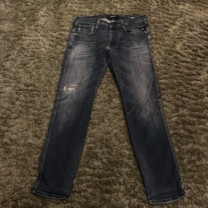 Replay Anbass blå jeans herr 32 - Säljer ett par Replay Anbass jeans i blå tvätt med slitningar och hål på vänster lår. Klassisk femficksmodell med raka ben och snygga detaljer på bakfickorna. Jeansen har en något faded look och är tillverkade i mjukt denimtyg.stretchande material på jeansen vilket gör dme väldigt sköna. 