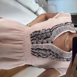 Ljusrosa blus med volanger - Supersöt ljusrosa blus med v-ringning och volanger framtill i svart och grått. Klänningen har korta ärmar och smock i midjan för en snygg passform. Materialet är lätt och luftigt, perfekt för varma dagar.