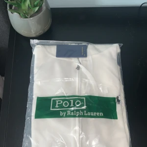 Vit hoodie från Polo Ralph Lauren M - Snygg vit hoodie från Polo Ralph Lauren i storlek M. Klassisk design med dragkedja framtill och broderad logga på bröstet. Mjuk bomullskvalitet och huva med snörning. Perfekt för en clean och stilren look.