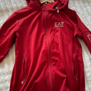 Röd EA7 vindjacka med huva - Snygg röd vindjacka från EA7 Emporio Armani med huva och silverfärgade logotyper på bröstet och ärmen. Jackan har dragkedja framtill, två fickor med dragkedja och ribbade muddar vid ärmslut. Perfekt för dig som gillar sportig och clean stil.