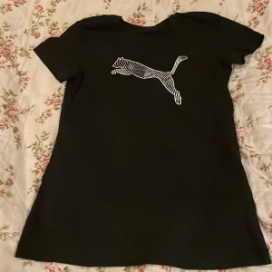 Svart Puma t-shirt med logga - Svart t-shirt från Puma i storlek XS med klassisk vit Puma-logga på bröstet. T-shirten har korta ärmar och är gjord i mjuk bomull som känns skön mot huden. Enkel och stilren design som funkar till många olika looks.