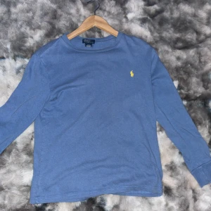 Blå långärmad tröja från Polo Ralph Lauren - Blå långärmad tröja från Polo Ralph Lauren med klassisk gul broderad logga på bröstet. Tröjan har rund hals och är gjord i mjuk bomull, perfekt för en chill och stilren look. Passar dig som gillar enkel design med en touch av lyx.