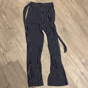 Randiga blå byxor med knytband - Snygga blåabyxor med vita vertikala ränder och knytband i midjan. Byxorna har en rak och lite vidare passform nertill, vilket ger en trendig look. Perfekta för dig som gillar stilrena och bekväma plagg med det lilla extra.