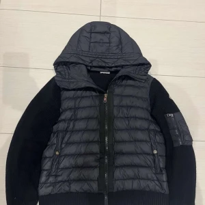 Svart Moncler pufferjacka med huva - Svart pufferjacka från Moncler med stickade ärmar och quiltad front. Jackan har en stor huva, dragkedja framtill och en ficka med dragkedja på ärmen. Materialmix av polyester och stickad ull ger en schysst kontrast. Perfekt för dig som gillar streetwear och exklusiva märken.